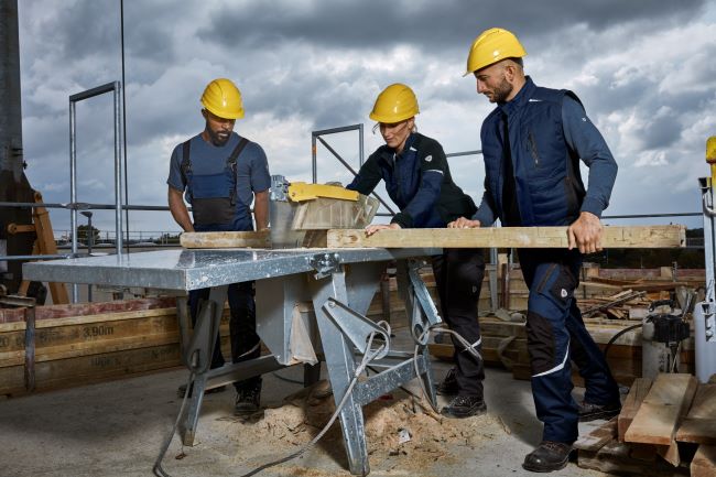 Zwei Dachdecker und ein Dachdecker tragen Arbeitskleidung und bearbeiten Holz auf einem Dach in Arbeitskleidung.